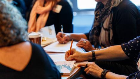 "A group in the middle of a discussion. They are sat at a table covered by paper and pens."